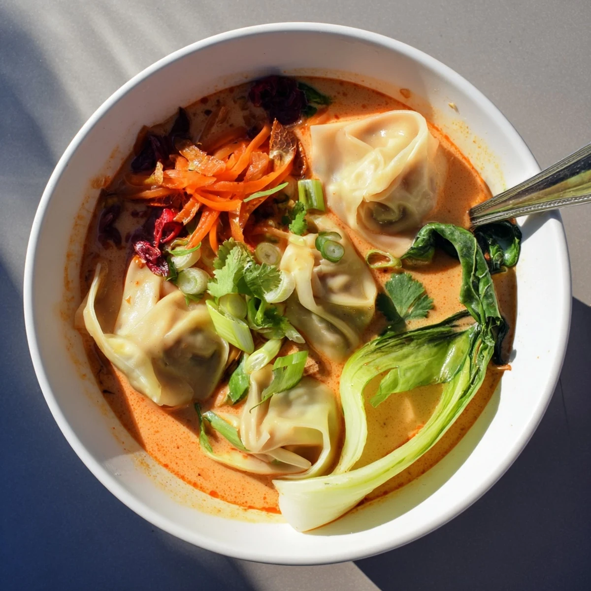 Bright red curry broth with plump wontons, shredded carrots, and wilted greens garnished with scallions and cilantro.  